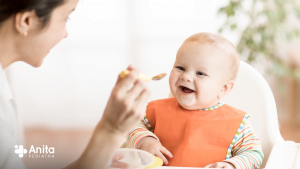 Uma imagem, contendo um bebê começando a introdução alimentar sentado em uma cadeira, uma colher e uma mulher