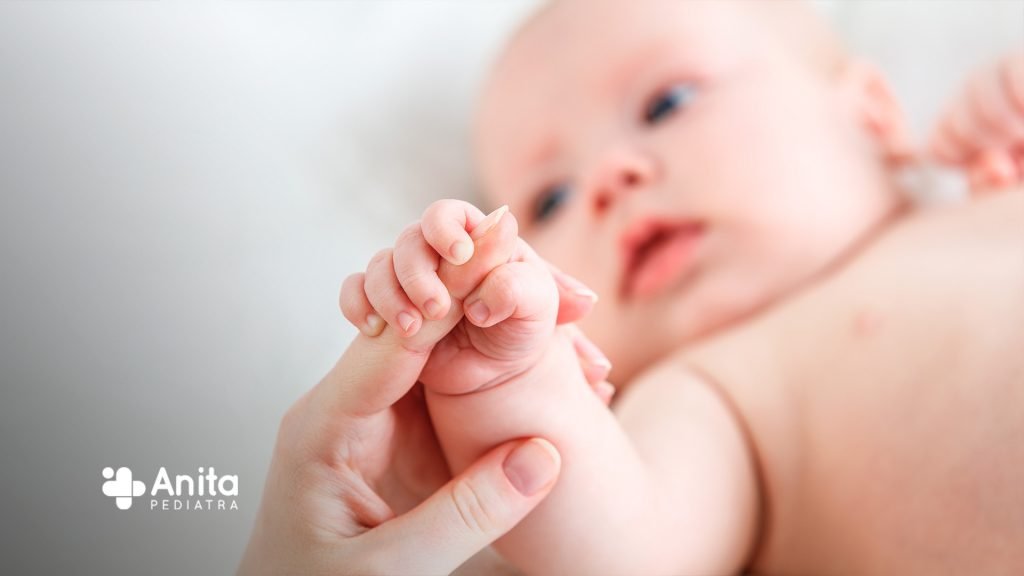 Uma imagem contendo um bebê no fundo, uma mão de adulto segurando-o para post sobre reflexos do recém-nascido.